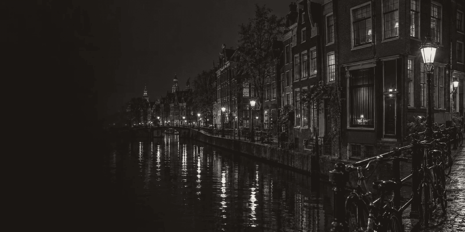 A black-and-white view along an Amsterdam canal at night, gabled brick houses, lamplight reflected in the water and bicycles chained to a bridge rail.