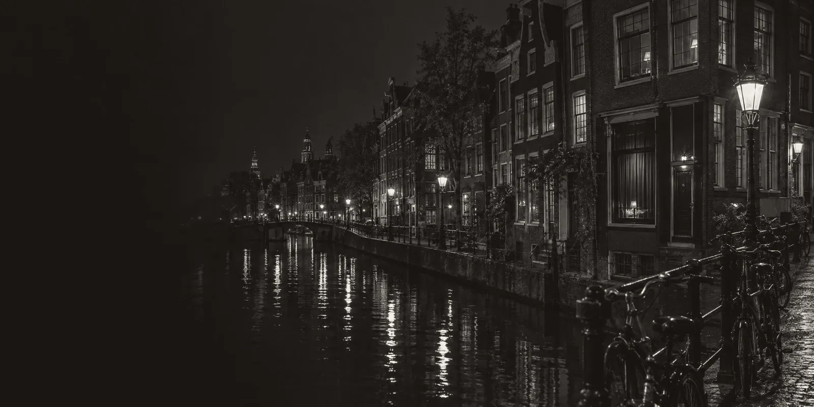 A black-and-white view along an Amsterdam canal at night, gabled brick houses, lamplight reflected in the water and bicycles chained to a bridge rail.