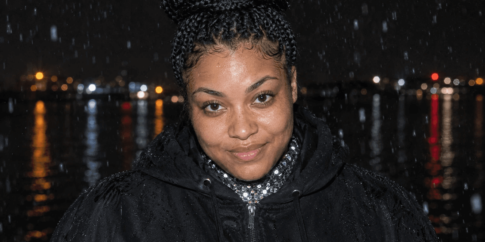 A young woman in her mid-twenties with long box braids wound into a high bun, broad strong shoulders, in a black hoodie zipped over the sequinned collar of a stage costume, caught by direct camera flash in the rain at night, chin lowered and a small shy smile, with blurred lights reflected in dark water behind her.
