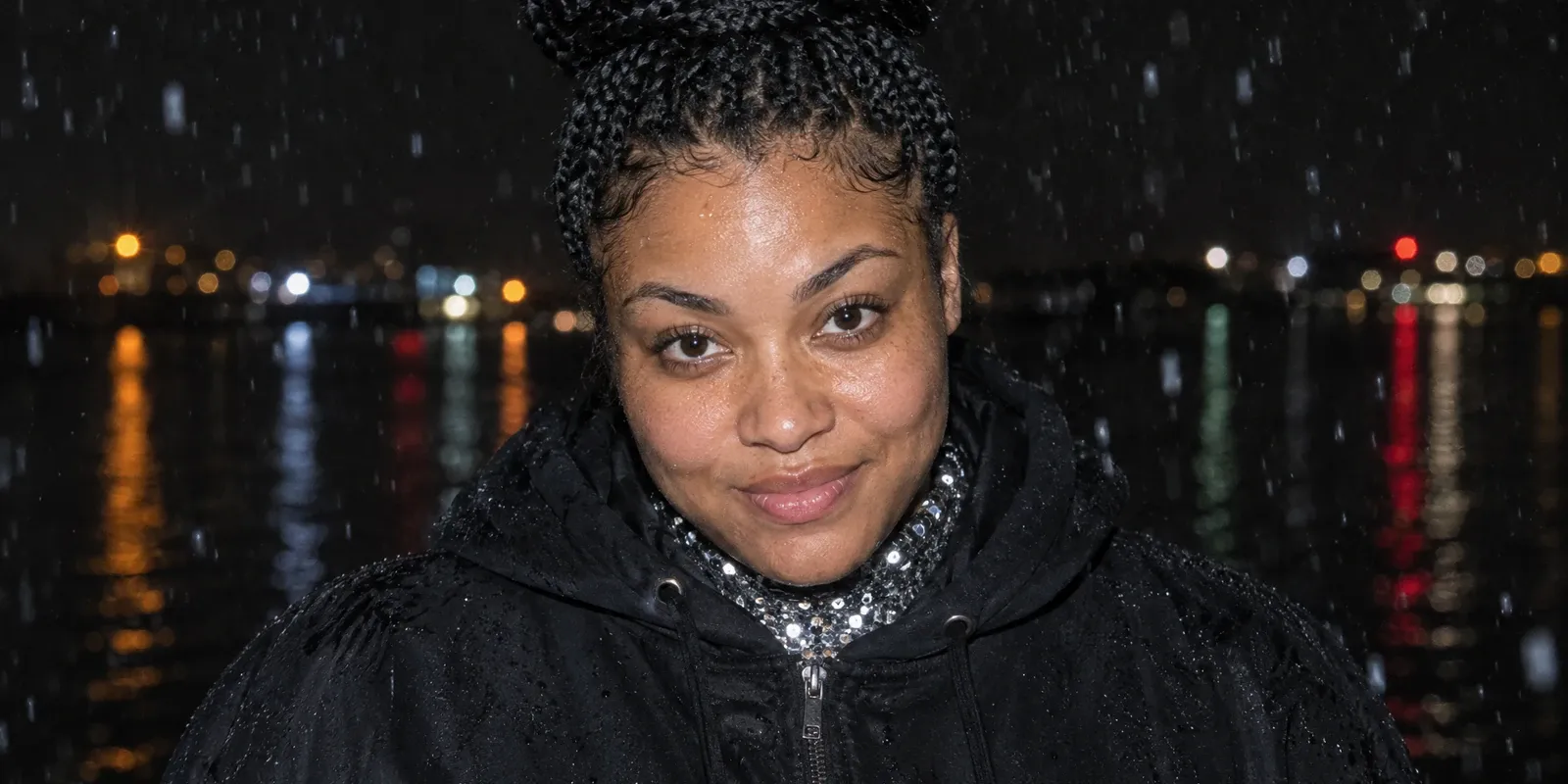A young woman in her mid-twenties with long box braids wound into a high bun, broad strong shoulders, in a black hoodie zipped over the sequinned collar of a stage costume, caught by direct camera flash in the rain at night, chin lowered and a small shy smile, with blurred lights reflected in dark water behind her.