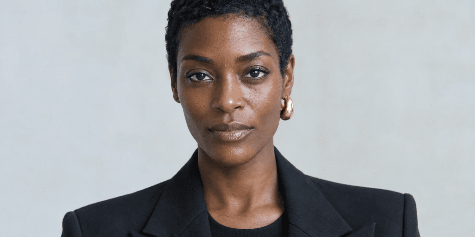 A woman in her early thirties with closely cropped hair and a gold hoop earring, in a black blazer over a black top, evenly lit in bright neutral light against a plain pale grey wall, her expression level.