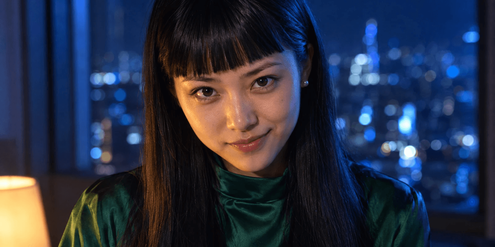 A young woman in her early twenties with long straight black hair and a heavy blunt fringe, in an emerald-green high-necked satin blouse, lit by cool blue window light and a small warm lamp from below, half-smiling, with blurred city lights behind the glass.