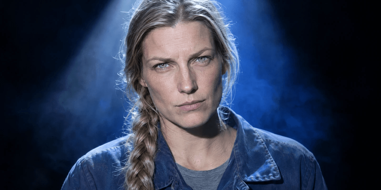 A tall woman in her mid-thirties with a long ash-blonde plait over one shoulder, in a faded blue work jacket over a grey T-shirt, lit from high above through white haze against black, eyes narrowed a little, unsmiling.