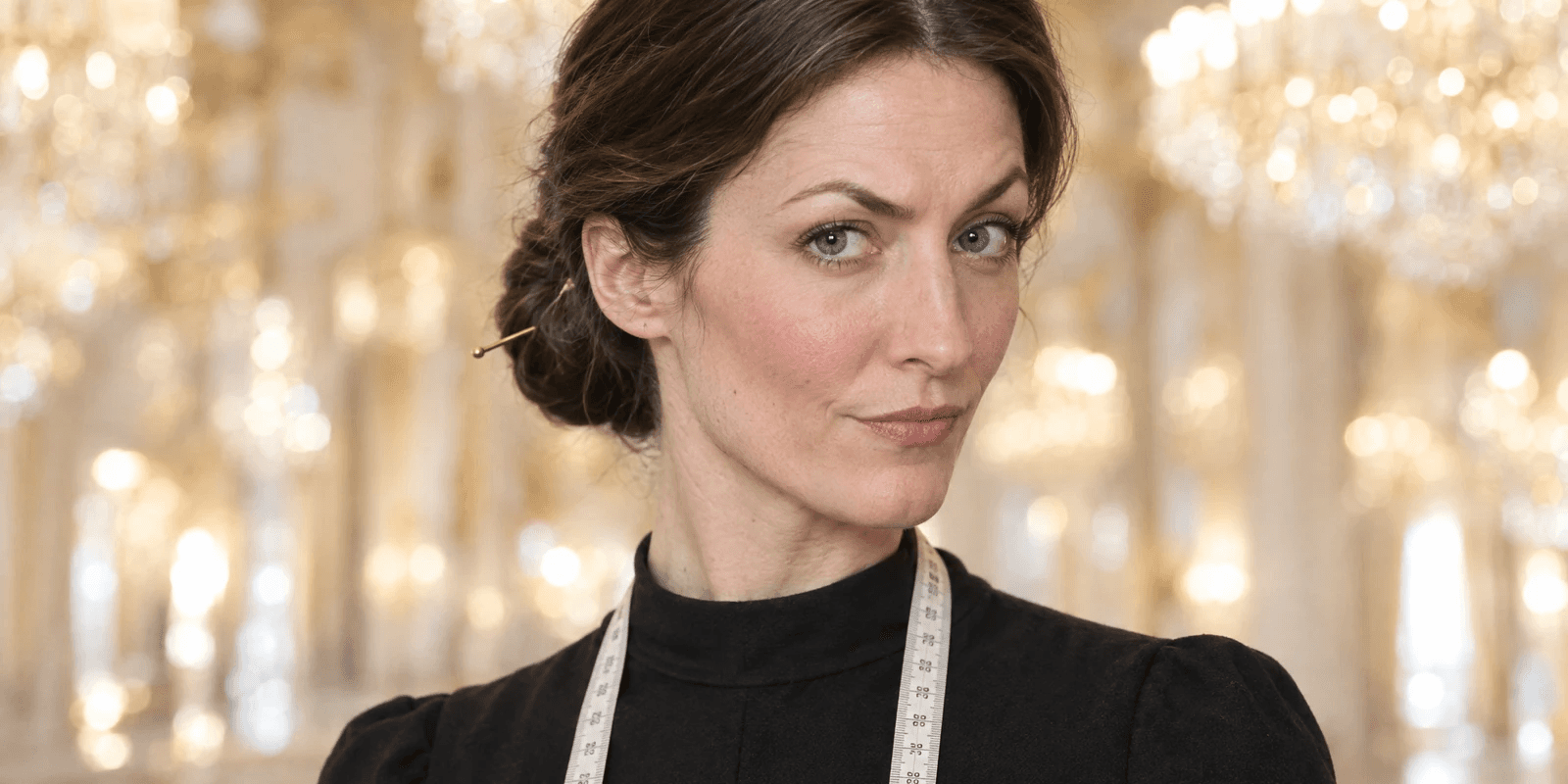 A woman in her late thirties with a strong aquiline nose, rosy fair skin and dark chestnut hair in a low knot with a needle pushed through it, a tape measure round the neck of her black dress, one eyebrow raised mid-verdict in bright soft chandelier light, with sparkling crystal and pale gold blurred behind her.