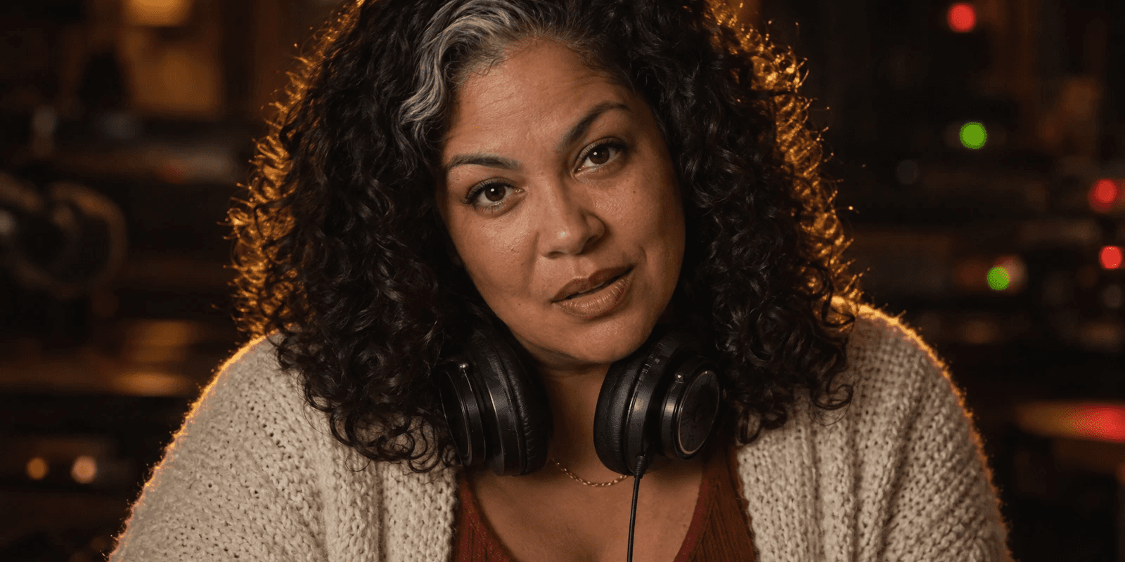 A woman in her early forties with thick dark curls and a grey streak at the front, headphones resting round her neck, in a cream knit cardigan, rim-lit in warm gold against a dark studio with small blurred indicator lights, caught mid-sentence.