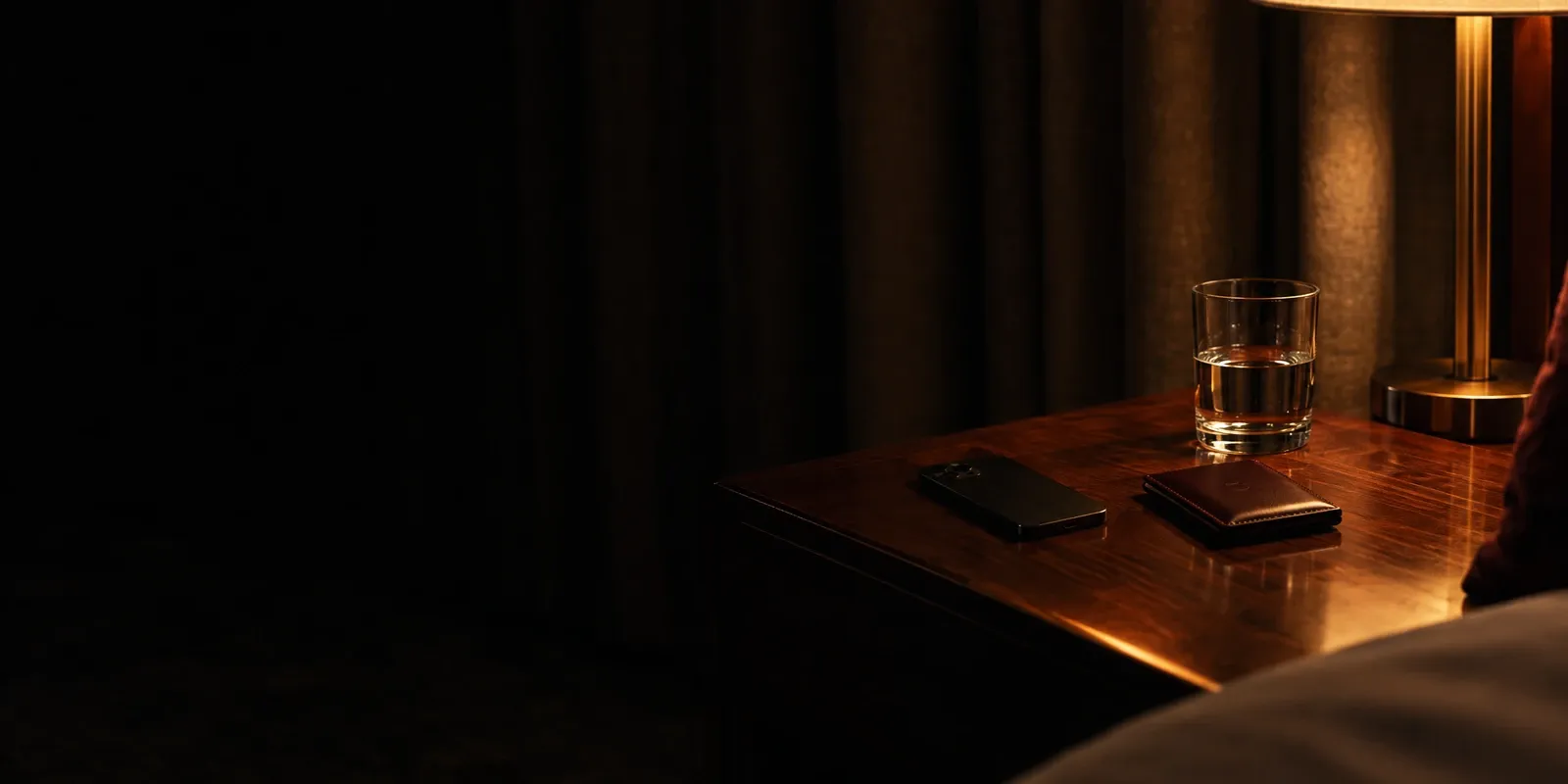 A smartphone lying face down beside a leather card wallet and a glass of water on a hotel nightstand, lit by one bedside lamp.